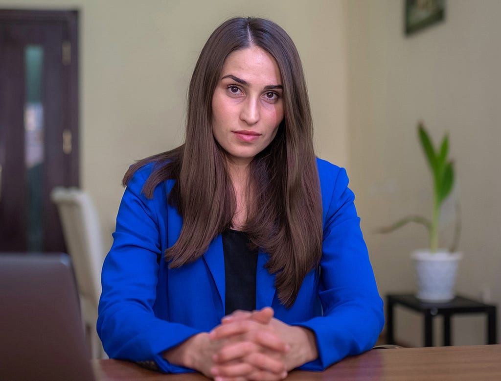 A woman with long brown hair wearing a blue blazer sits in an office setting looking at the camera.