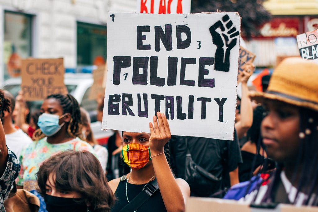 An image of a crowd of protesters. In the center, a woman with brown skin and a colorful face masks holds up a white sign with “End Police Brutality” in black writing and a Black Power fist.