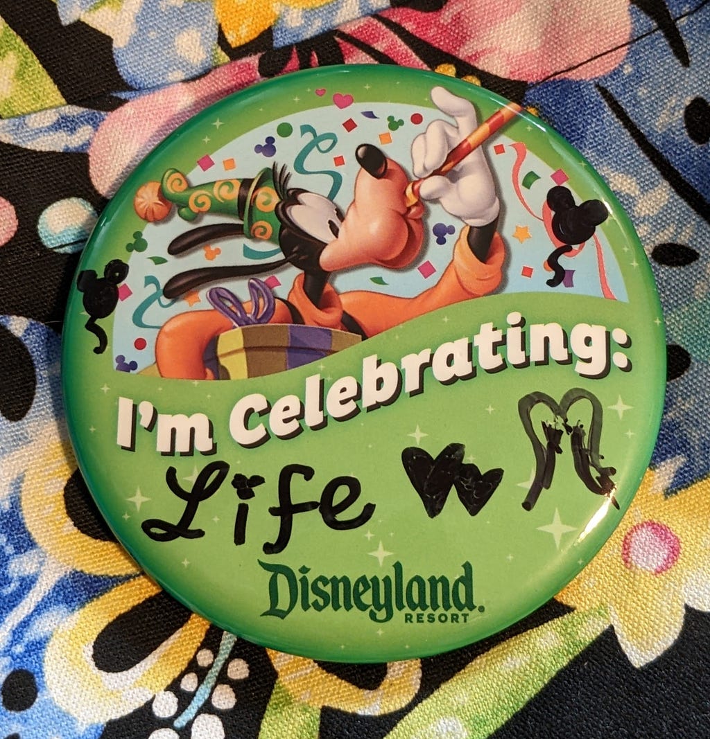 A Disneyland pin saying “I’m celebrating:” with marking writing out “Life” followed by two hearts and really messy angel wings