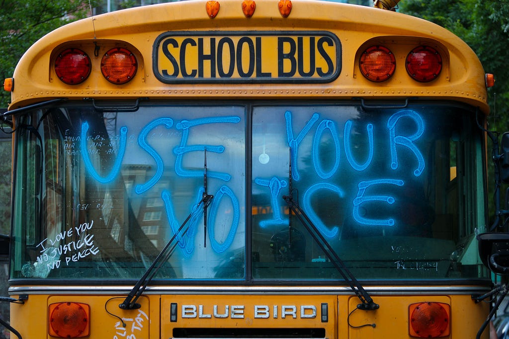 Close up of the front windshield of a big, yellow school bus. On the window, in big, bright, blue spray paint, the words “Use Your Voice” and smaller, in white marker, the words, stacked: “I Love You” “No Justice” “No Peace.” Below the windshield, the bus logo reads, “Blue Bird” in metal letters,  and above the windshield are the words, “School Bus” in black.
