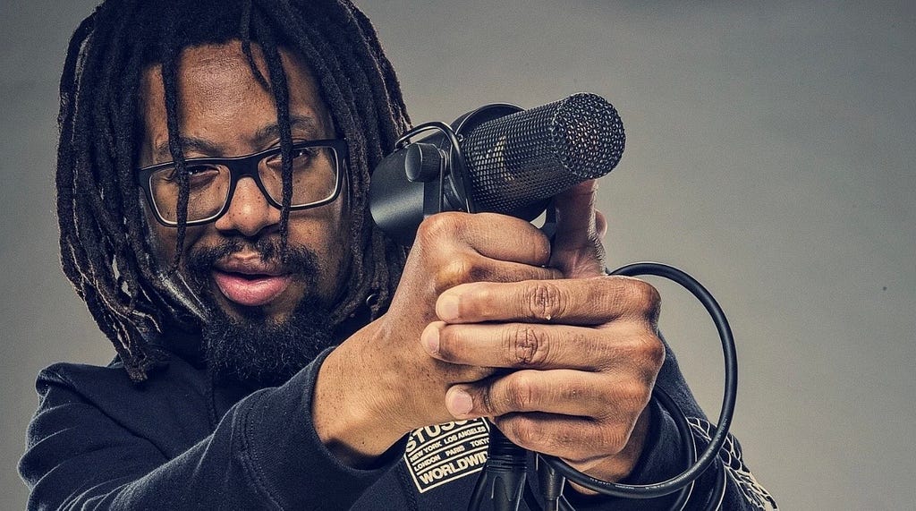 Boston rapper Mr. Lif aiming a microphone like a gun. Caption says “his most powerful weapon against the world.”
