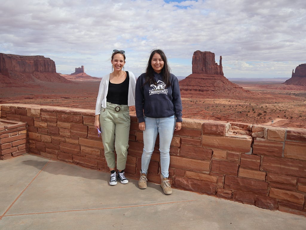 Two women standing together against a low red-brick wall, smiling for the camera, wearing casual clothes. In the background are giant red monolithic rocks in a desert landscape.