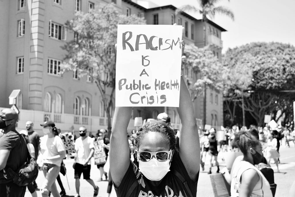 Black woman wearing a surgical mask faces the camera holding protest placard that reads: RACISM IS A Public Health Crisis.”