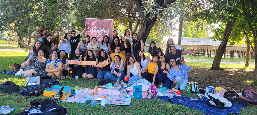 Grupo de mujeres que asistieron a la junta presencial de +Mujeres en UX Chile 2023. Todas están frente al pendón con el logo de la organización, una fila de pie y otra fila sentada. Algunas saludan a la cámara y otras sonríen.