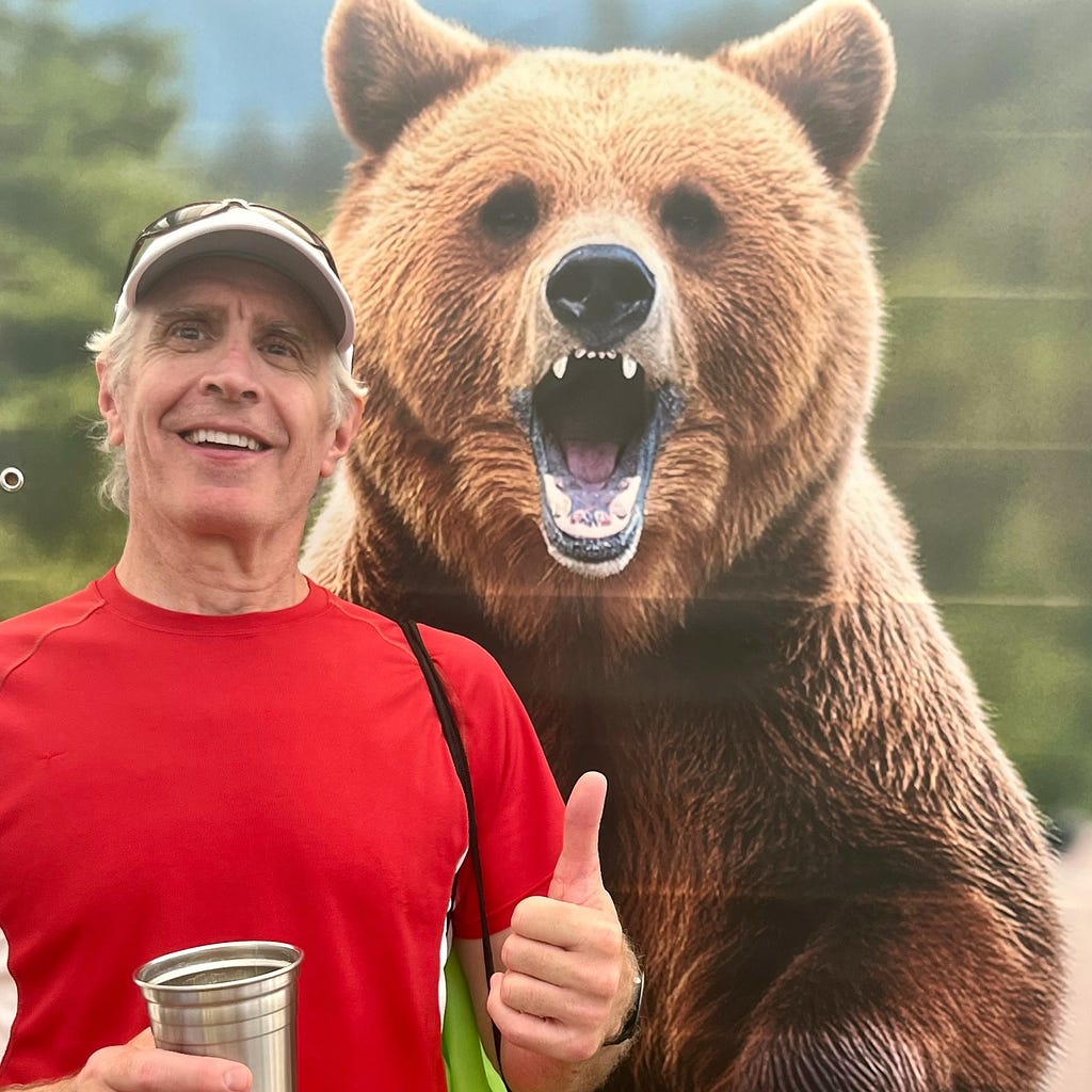 An older man wearing a smile and a red t-shirt stands in front of a grizzly bear poster