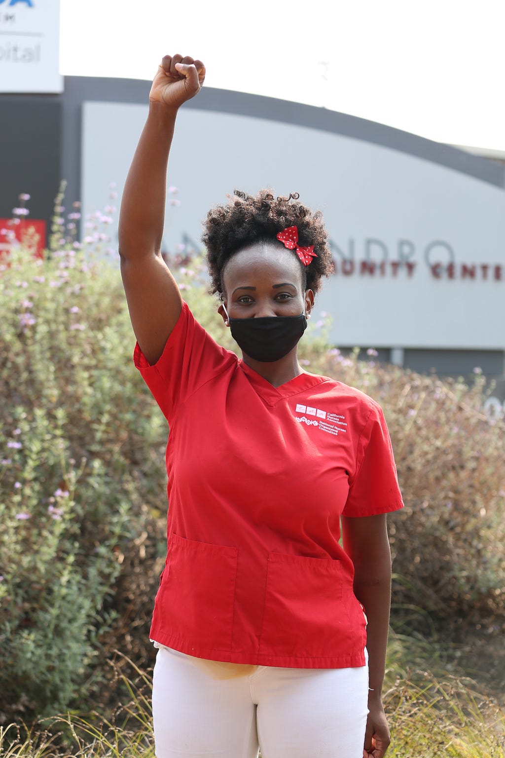 Image of Mawata Kamara — a Black nurse wearing red scrubs, a black face mask, and a red bow in her hair — with her first raised in the air.
