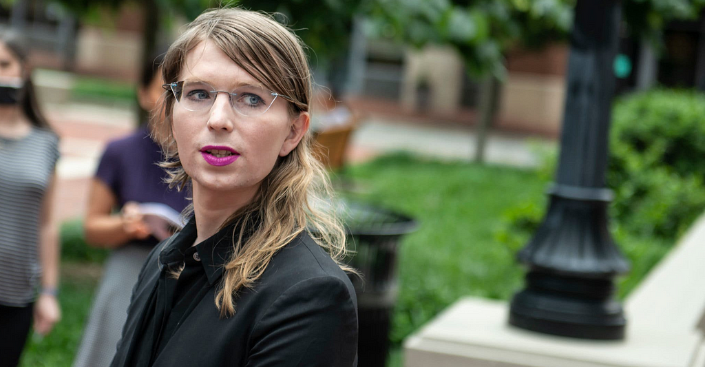 A photo of Chelsea Manning, a white woman with blonde hair past her shoulders, pink lipstick, glasses, and a black suit, looking to the right.