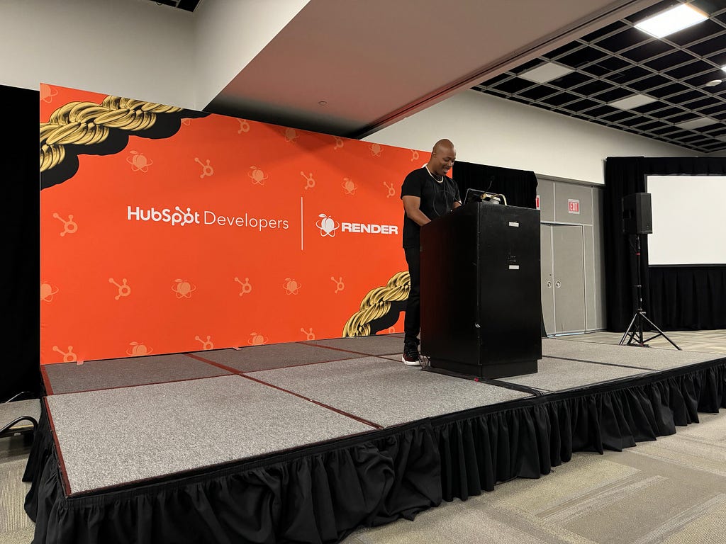 Mark (Techson) Thompson giving an early morning presentation on web development on a small stage in front of a large orange and gold backdrop.