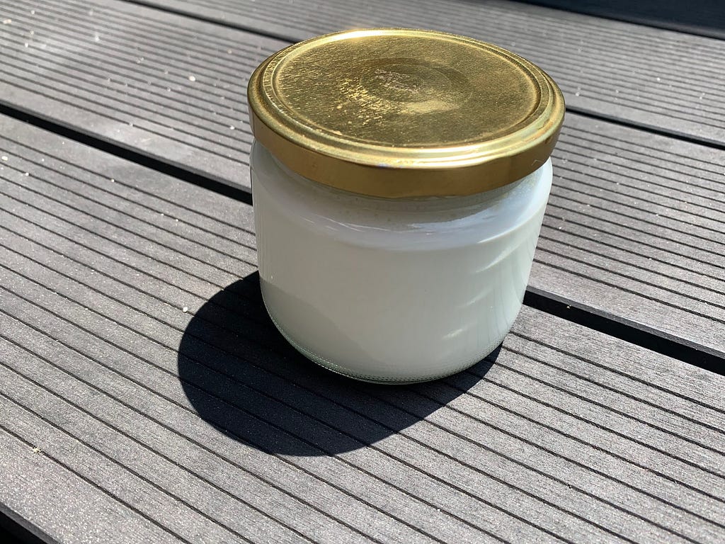 A jar of homemade yogurt with a gold lid sitting on a wooden deck in the garden on a sunny day.