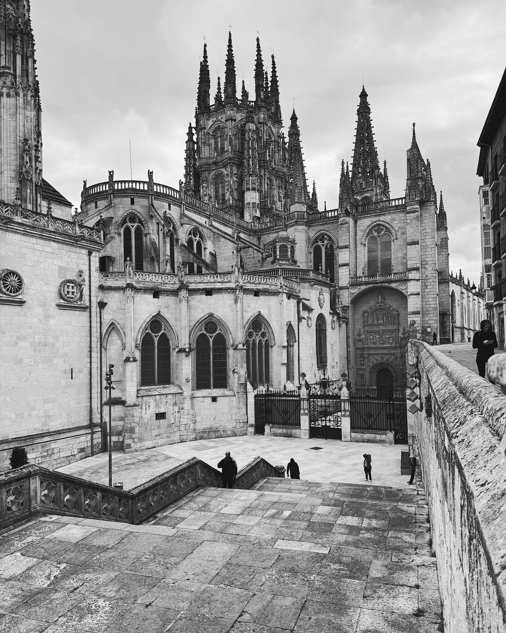 Black and white close-up photo of Gothic architecture of large Burgos Cathedral. We are looking at the side entrance with less elaborate ornamentation. The spires rise up in the background.