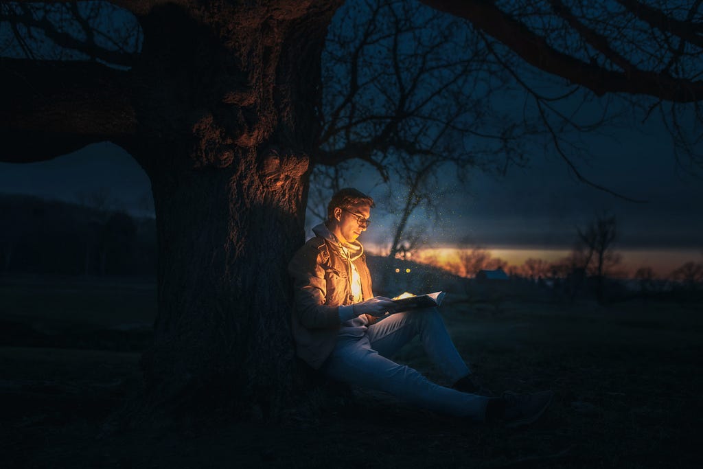 A man sitting against a tree at night. He has an open book in his lap and light is eminating from it, illuminating his chest and face.