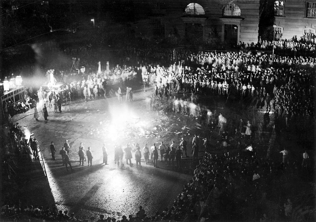 A black and white photo showing a large crowd of people surrounding paper materials on fire.