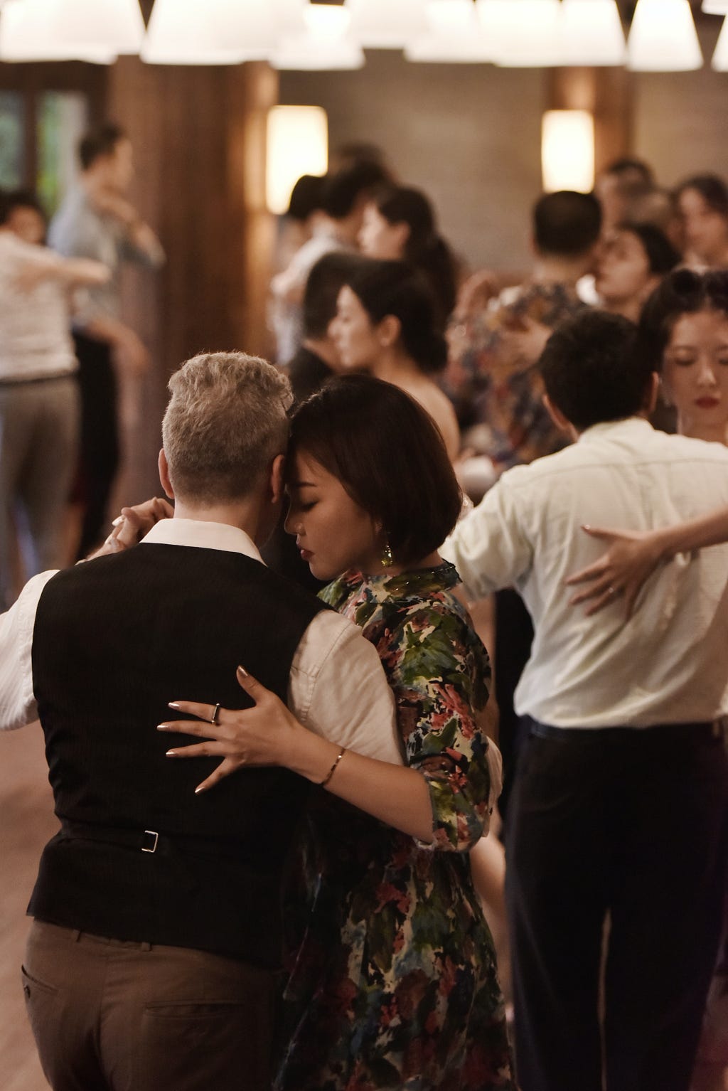 A group of people dance in a room. They are dancing in couples, each couple in a close embrace.