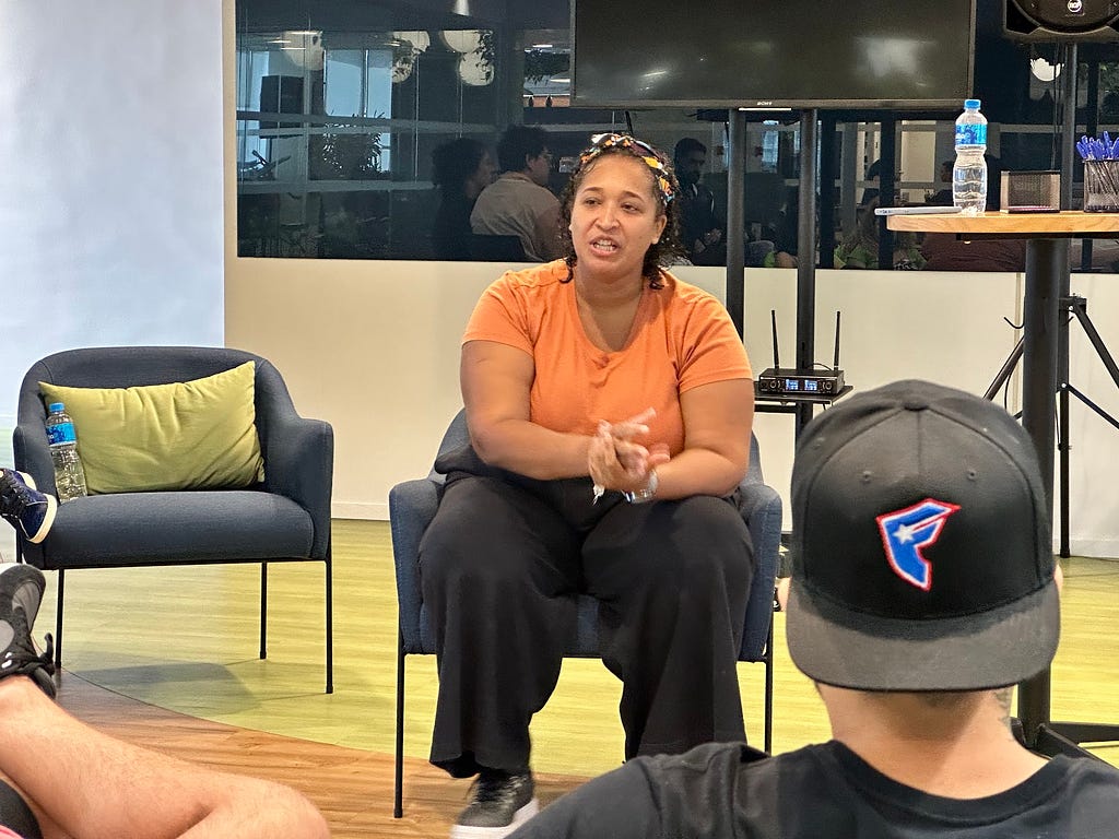 Priscila Cardoso, uma mulher negra, alta e grande, sentada em uma cadeira enquanto fala para nossa equipe, que assiste seu painel.