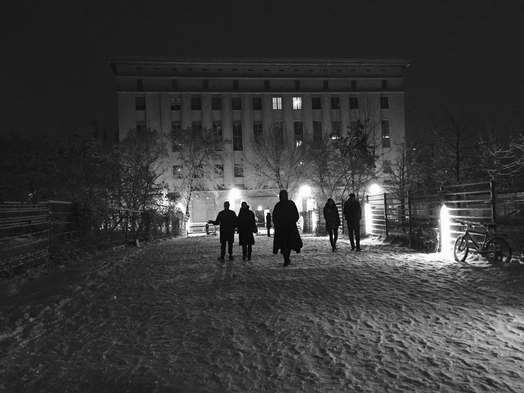 A black and white photograph of a warehouse nightclub in the snow with some siloutetted figures approaching the building.