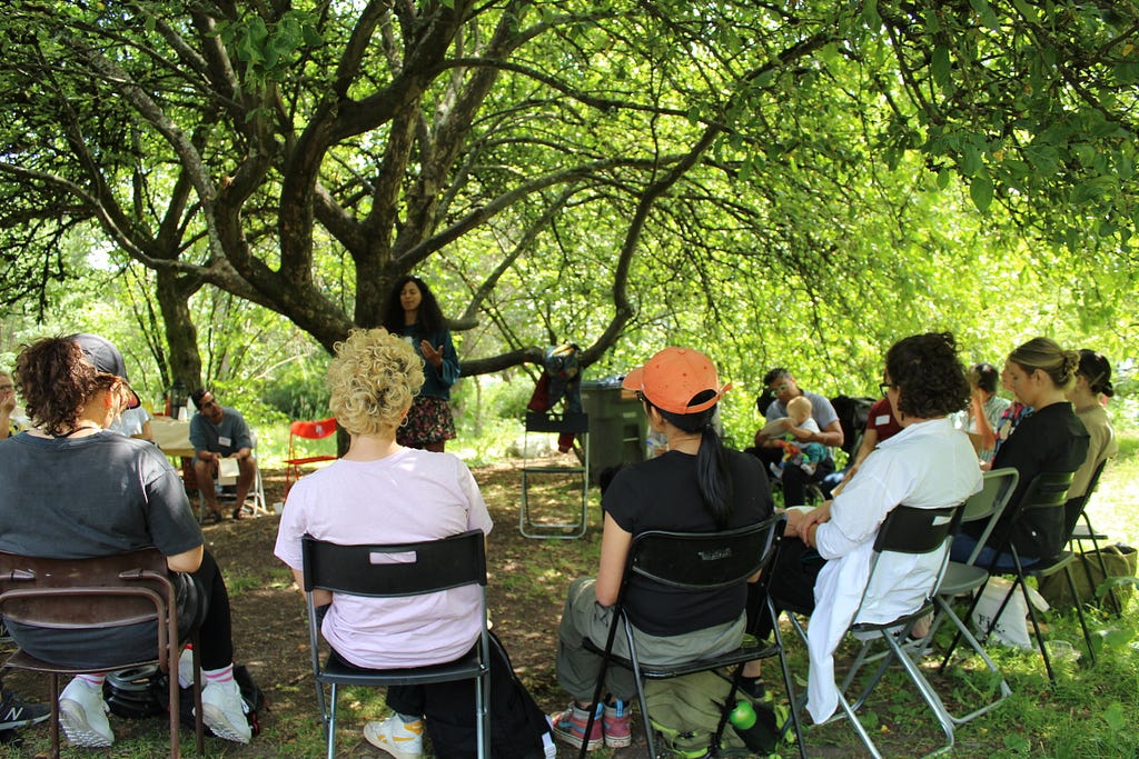 A group of about 20 people are seated on folding chairs under a canopy of leafy trees listening to one person who’s standing with eyes closed and hands outstretched.