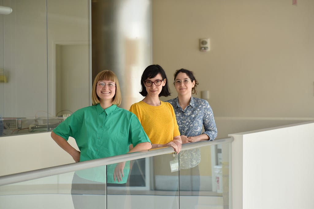 Taylor Baum, Josefina Correa Menéndez and Karla Alejandra Montejo stand along a glass railing in a white-hued corridor of MIT’s Building 46.