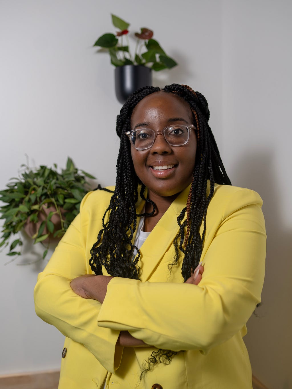 A self portrait image of Kara Branch smiling. She is folding her arms and wearing a yellow dress suit with brown buttons. She has clear glasses on and light brown eyeshadow. Her hair is in braids pulled back into a half up half down hairstyle. Her braids have brown highlights.