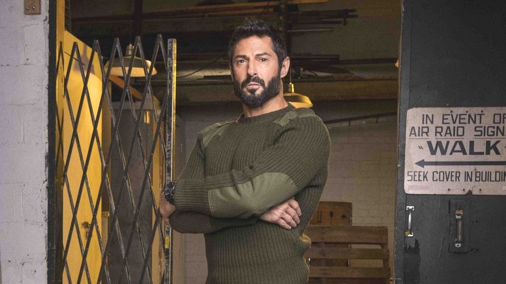 Rudy Reyes with arms crossed in an olive long-sleeved shirt in a warehouse