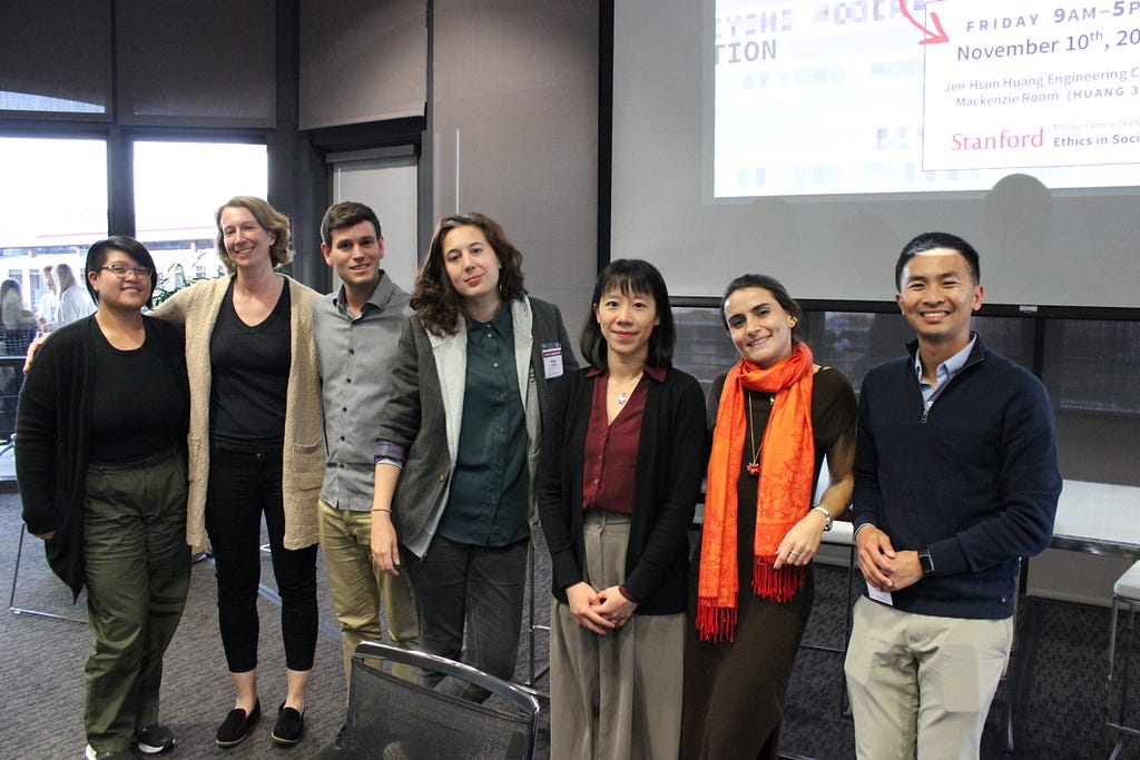 Seven professionally dressed people standing inside and looking at the camera and smiling.