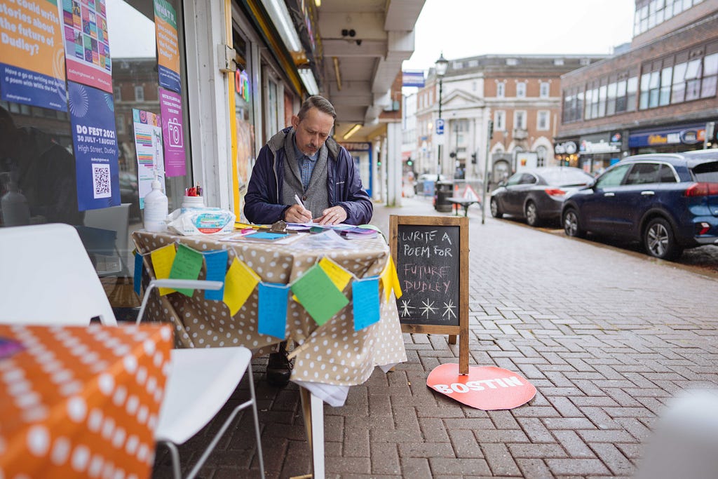 Photo of Rick, a middle aged white man, sitting on Dudley High Street at a small table with a brown spotty tablecloth, leaning over writing. To the left are windows of a High Street retail unit which is home to CoLab Dudley, to the right are parked cars and traffic moving along the street. A small chalkboard next to Rick invites people to write a poem for Dudley.