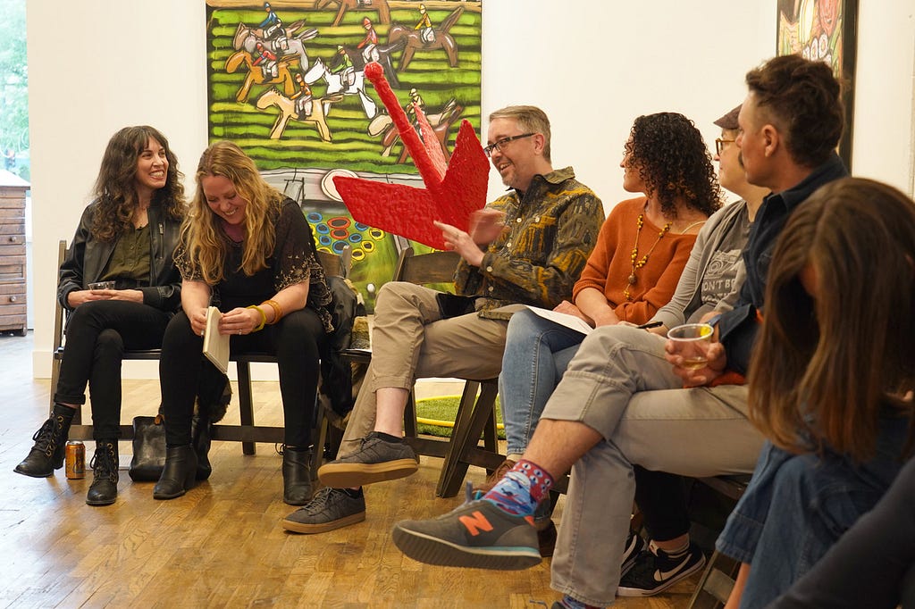 A group of six men and women sit in a semi-circle talking — behind them is a green painting with animal figures and red arrow-shaped sculpture.