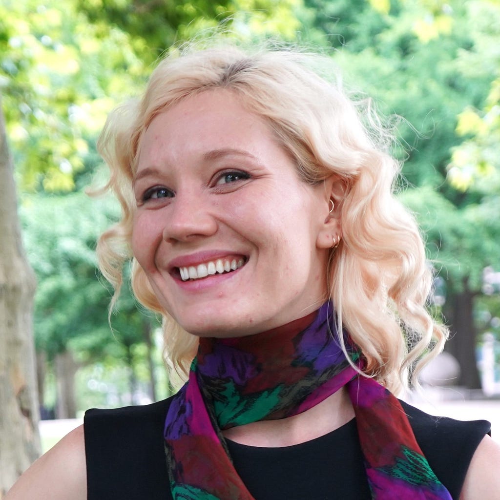 A white woman in her 30s with blond curly hair smiling and wearing a black shirt with a colorful scarf poses in a park.
