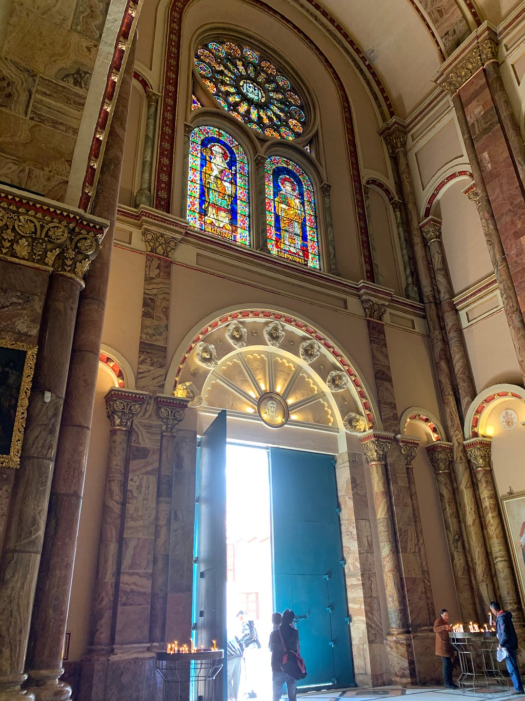 Interior of Cuenca’s Cathedral. Red, blue, pink and white stain glass windows of two individuals sit above the side entrance.