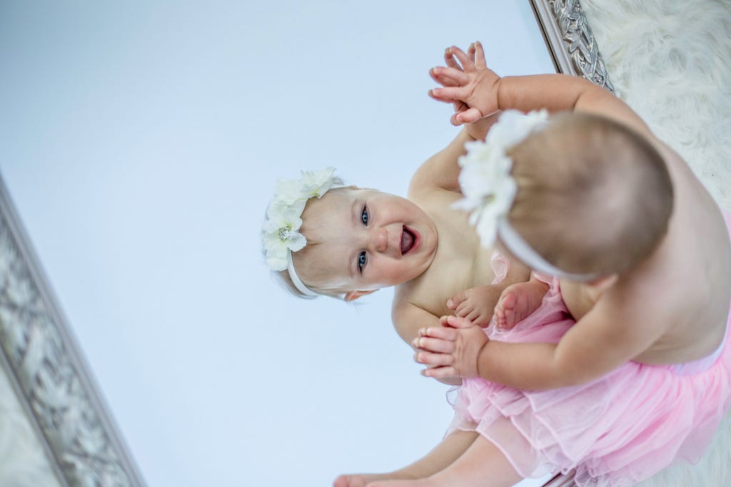 A baby dressed in a pink tutu and a flowery head-band delighting in her reflection in the mirror.