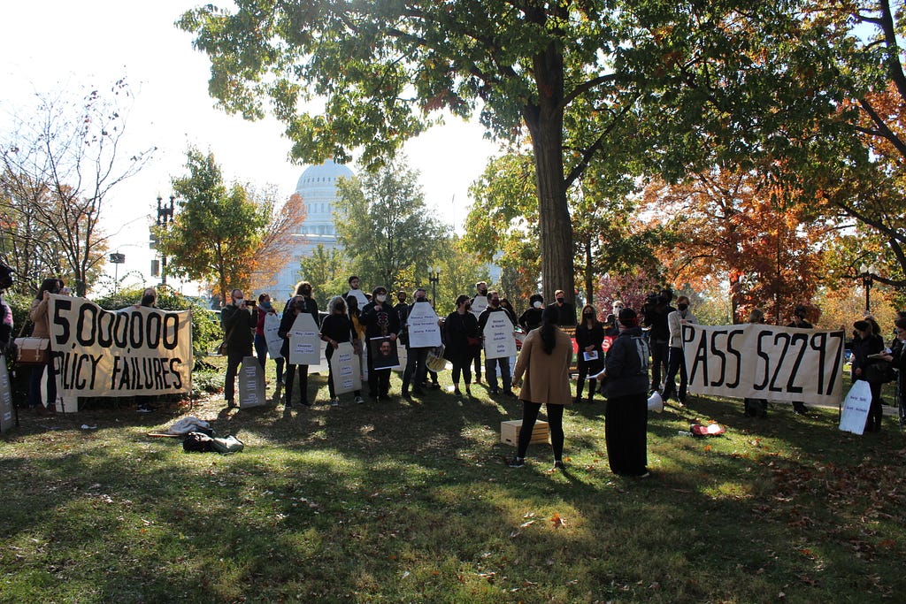 A group of people are gathered outside in a large grassy area with the US Capitol building in the background, underneath a tree. There are smaller groups of people within the larger group that are holding banners that read, “5,000,000 Policy Failures” and “Pass 52297.” Others in the group are holding signs. There are two people standing in front of the group, facing them and speaking.