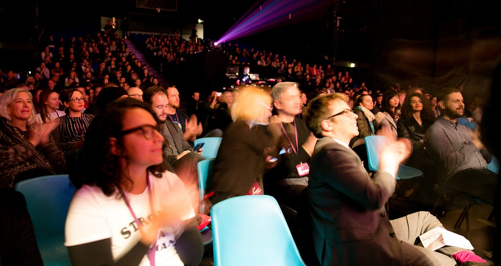 An audience of Interaction Awards attendees applaud. They are seated in a theatre and looking at the stage which is out of frame to the right.