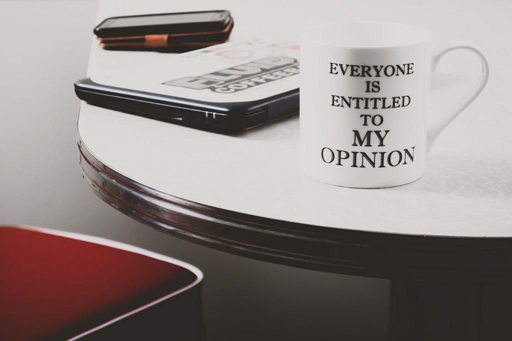 Coffee mug on a table printed with the slogan “Everyone is entitled to my opinion”