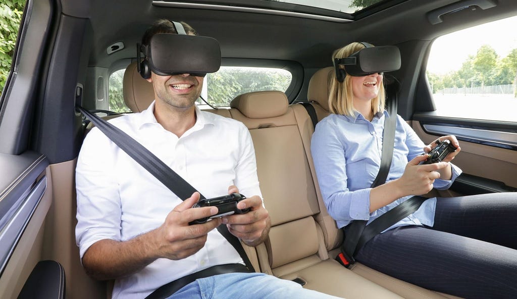 Photo of two people in the rear of a car, wearing virtual reality headsets and holding game controllers