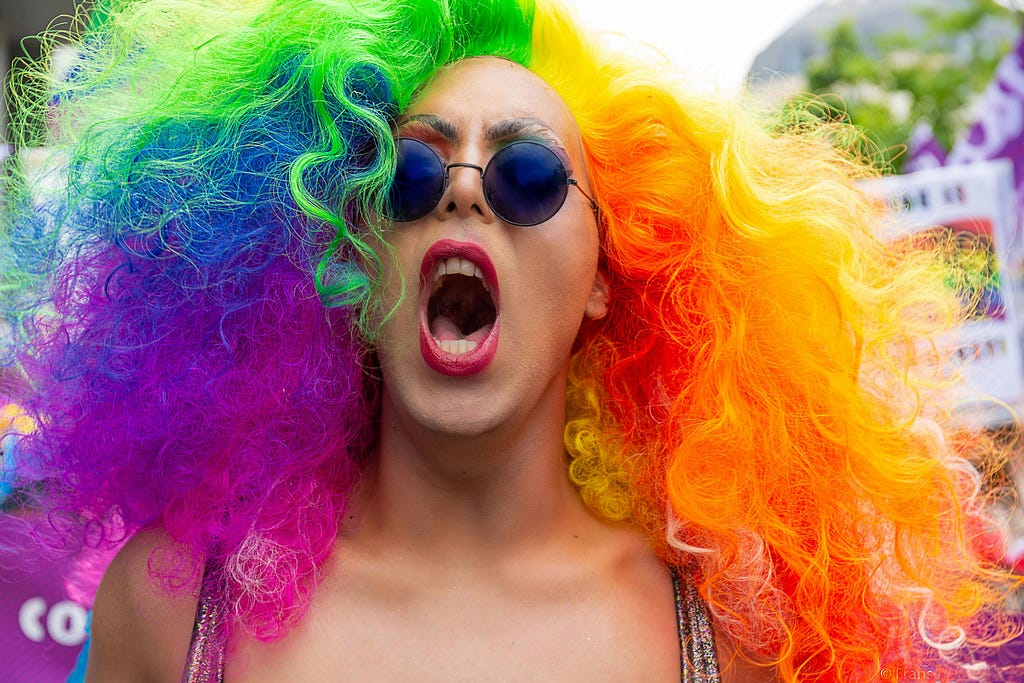 Headshot of person with long frizzy rainbow hair and eyeshadow, round sunglasses, and red lipstick yelling at the camera