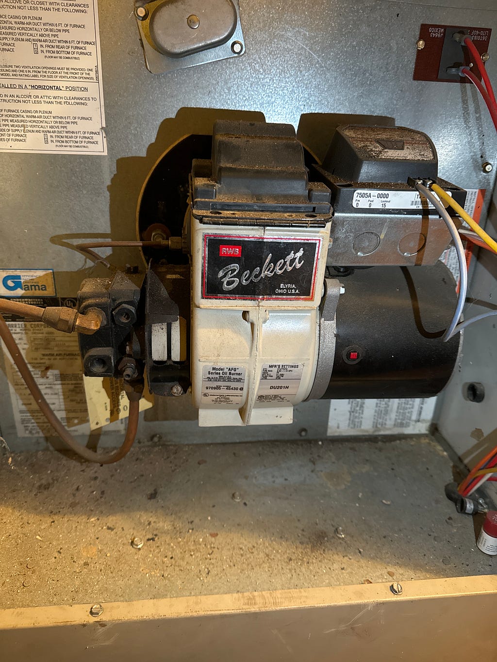 A close-up photograph of a Beckett oil burner mounted inside a furnace, showing warning labels, control switches, and wiring. The unit appears well-used with some dust accumulation, indicative of regular operation.