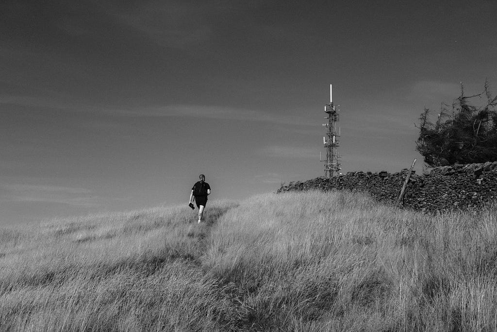 A black-and-white photo of Tom Illsley walking up a hill towards a telecommunications tower, image by Ian Howorth