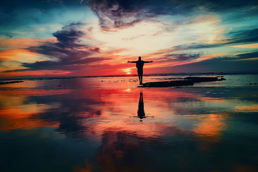Person standing on the beach with arms wide open in looking out onto a multicolored sunset.