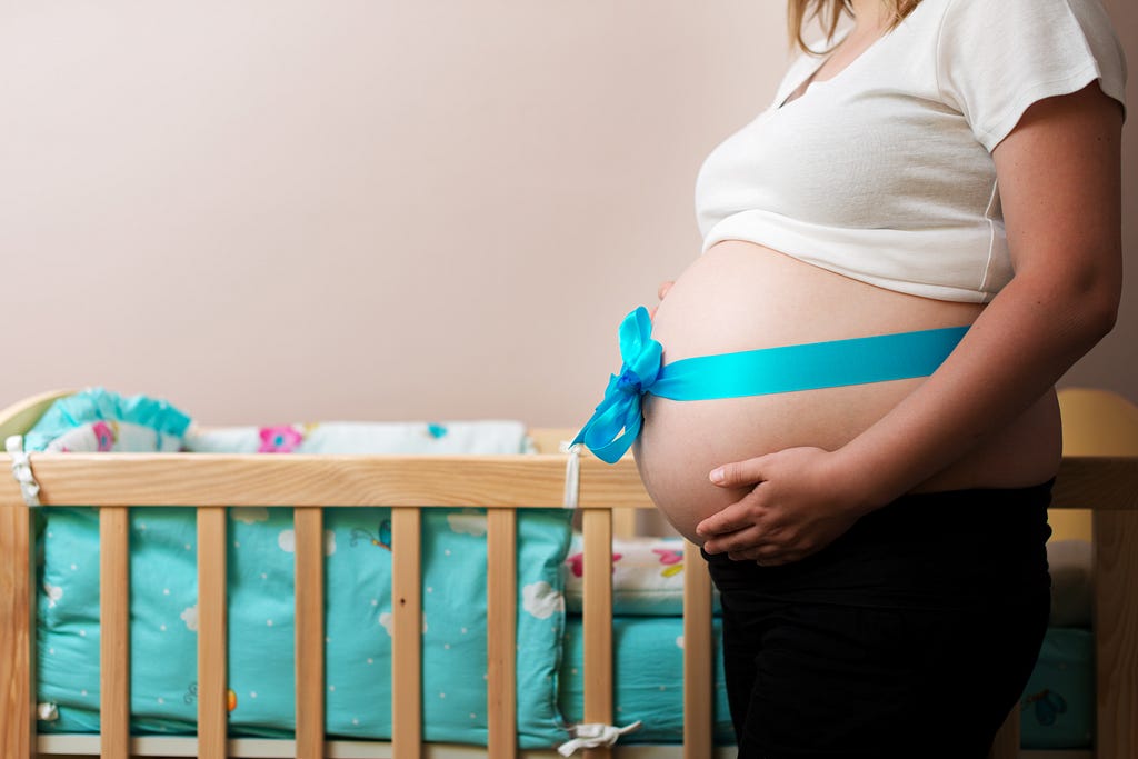 Pregnant belly wrapped in ribbon with a bow, in front of empty cot