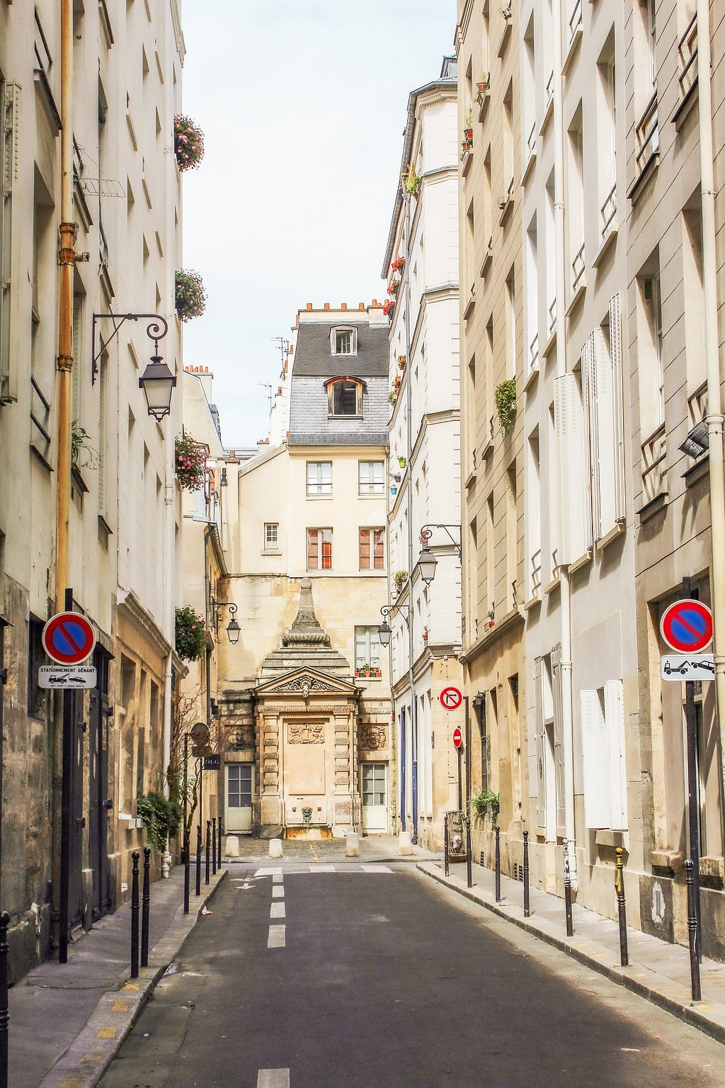 narrow street in paris with tall buildings, lantern and fountain at the end.