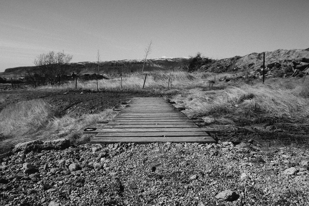 Gravel path leads to worn wooden planks extending into a rocky dry grass landscape without trees