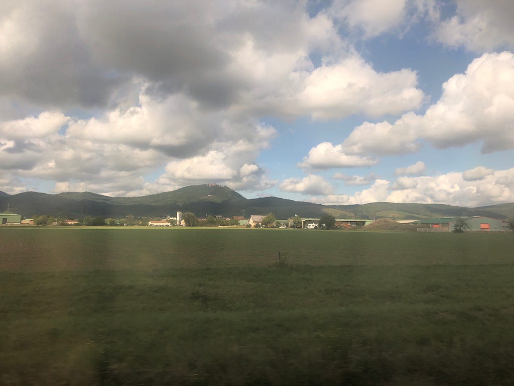 (Picture taken through a train window) White clouds and sun shining through them. Mountains in the background, a few buildings, and fields in the foreground.