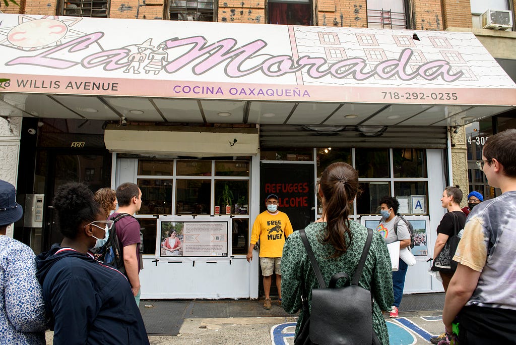 A group listens to a man in front of two banners, visible on the storefront of the restaurant La Morada