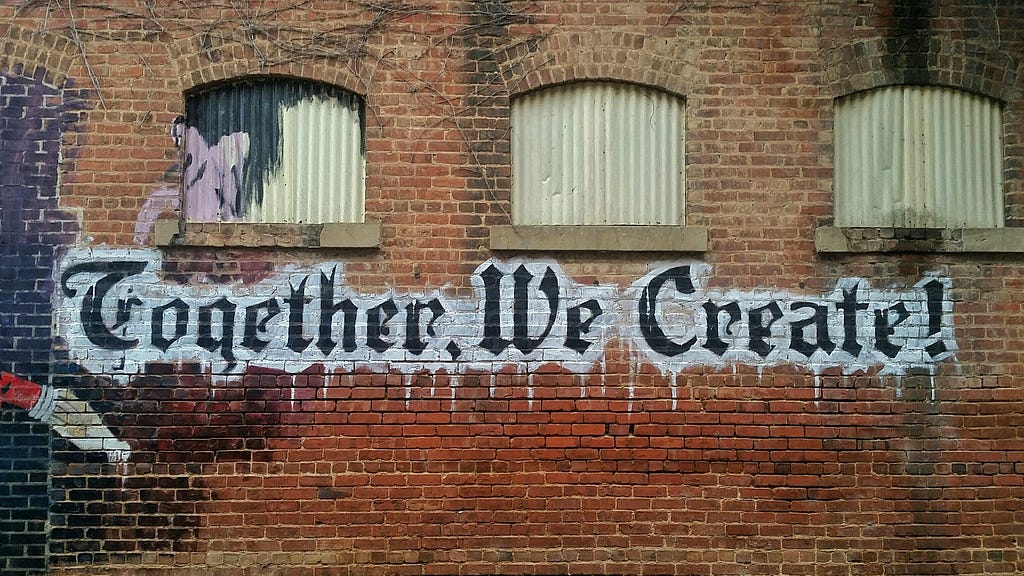 “Together, We Create!” spray painted in black lettering on an urban brick wall with shuttered windows.