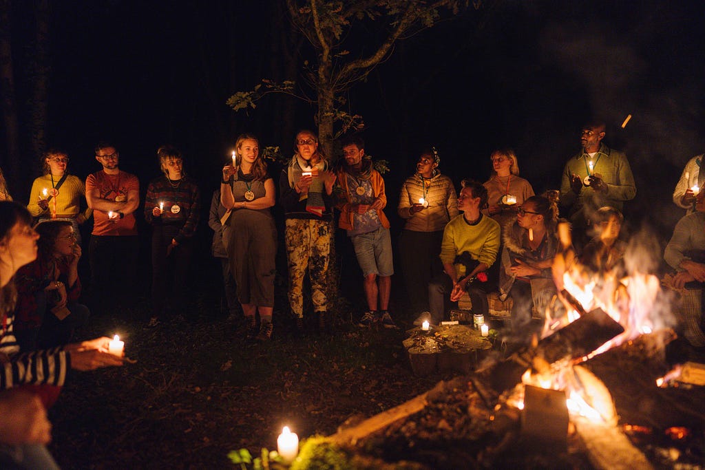 A group of people holding candles stand around a fire at night.