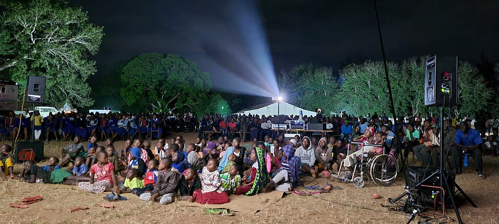 Hundreds of people in Kenya sit on the ground with projector light shining behind them as they watch a mobile cinema screening of “The Elephant Queen.”