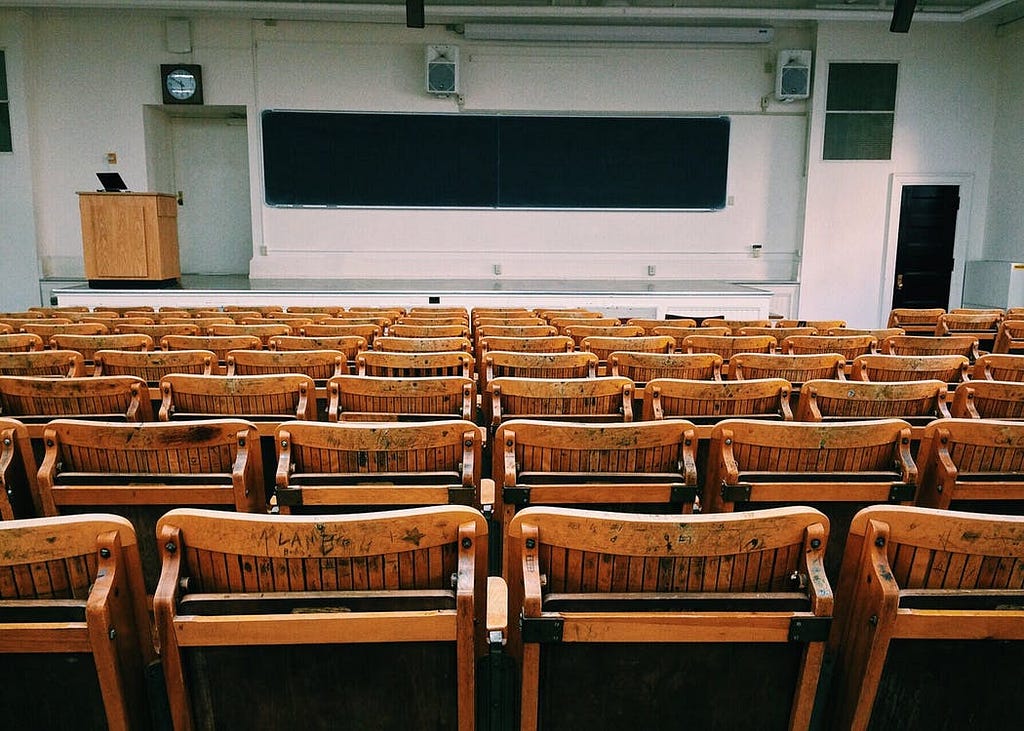 Empty classroom with brown and black wooden chairs inside large room with podium and no people