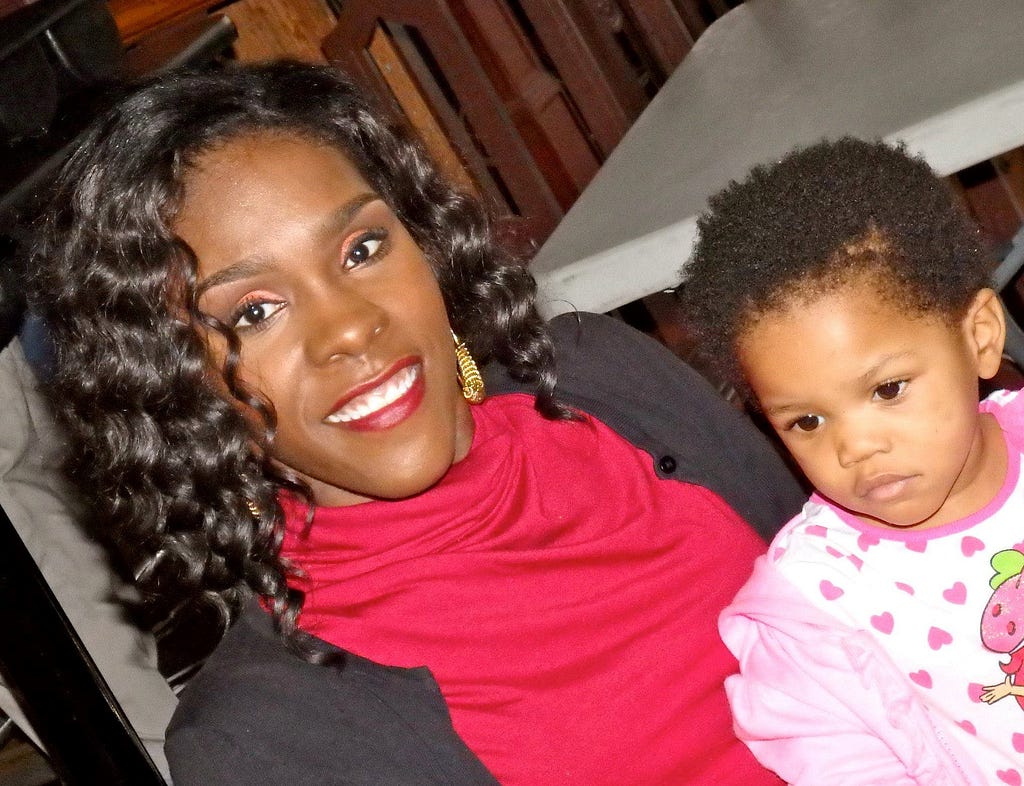 A picture of Tameka Citchen-Spruce. She has wavy hair and is wearing a red turtleneck and black cardigan. Her baby daughter is sitting on her lap and is wearing pink heart-print shirt.