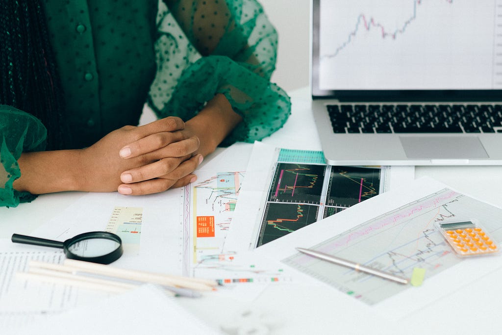 Hands clasped on a desk, cluttered with notes and graphs on paper, placed beside a laptop.