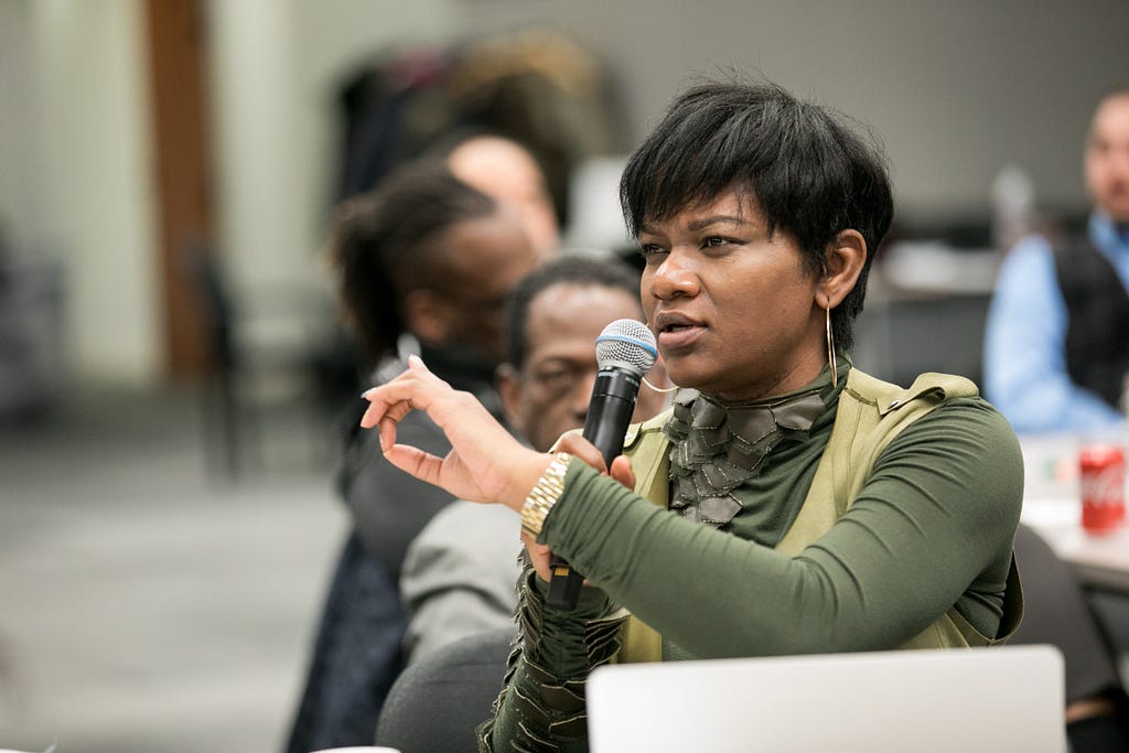 Nakia speaks into a microphone during an Institute training session. Behind her, Fellows Art Sims and Robert Green take notes