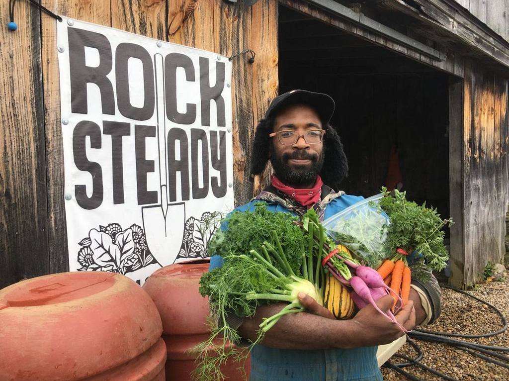 Farmer holds a bundle of carrots and other seasonal produce in front of a barn with a Rock Steady logo